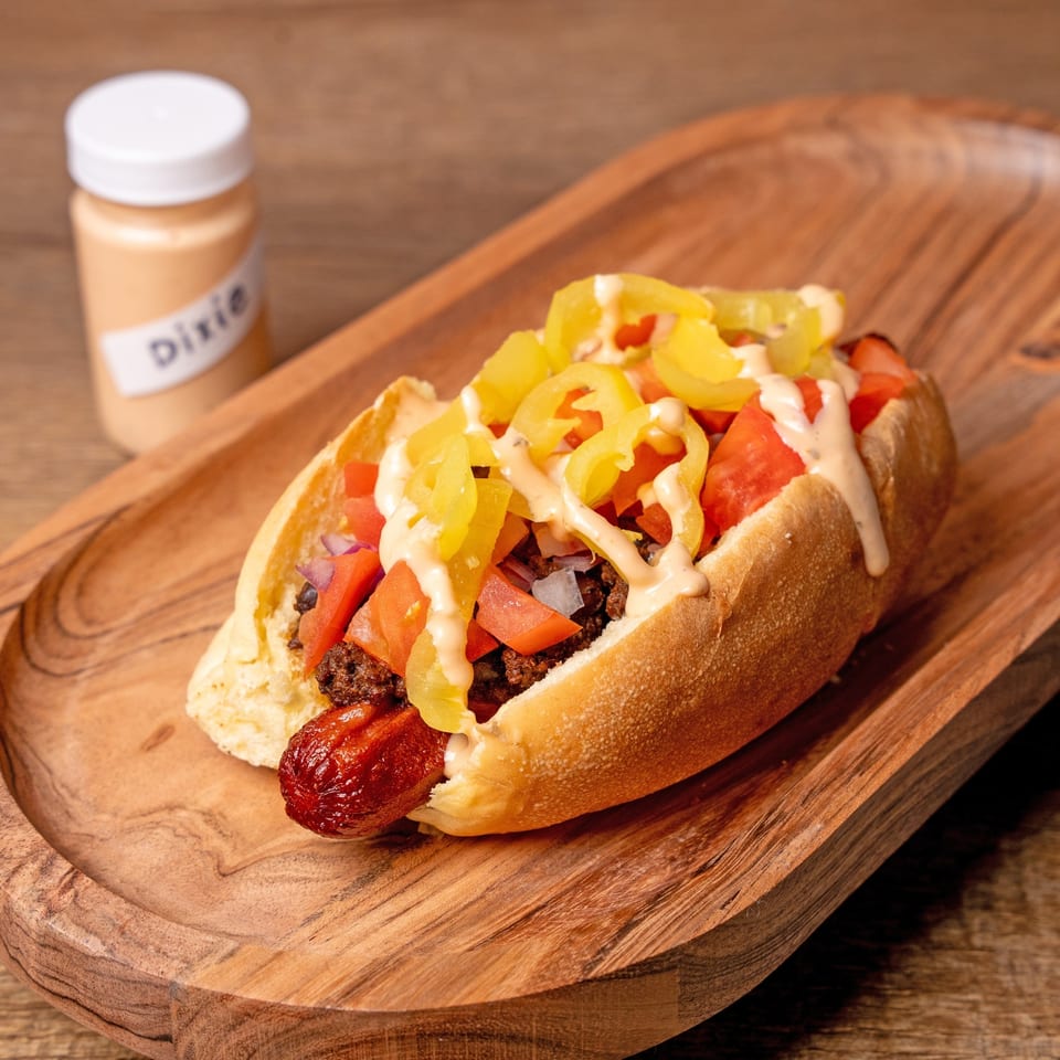 A hot dog with chili, diced tomatoes, yellow peppers, and sauce on a wooden tray.