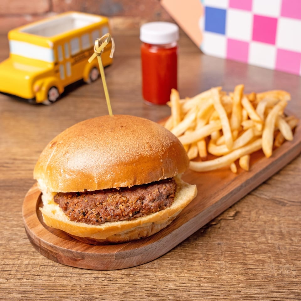 A burger and fries on a wooden board, with a toy school bus and ketchup bottle in the background.